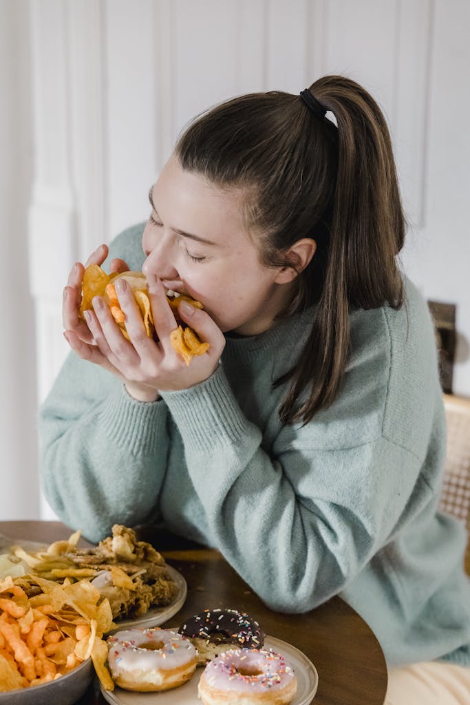 Hungry female in casual clothes eating crispy chips from hands while sitting at table with assorted junk food at home