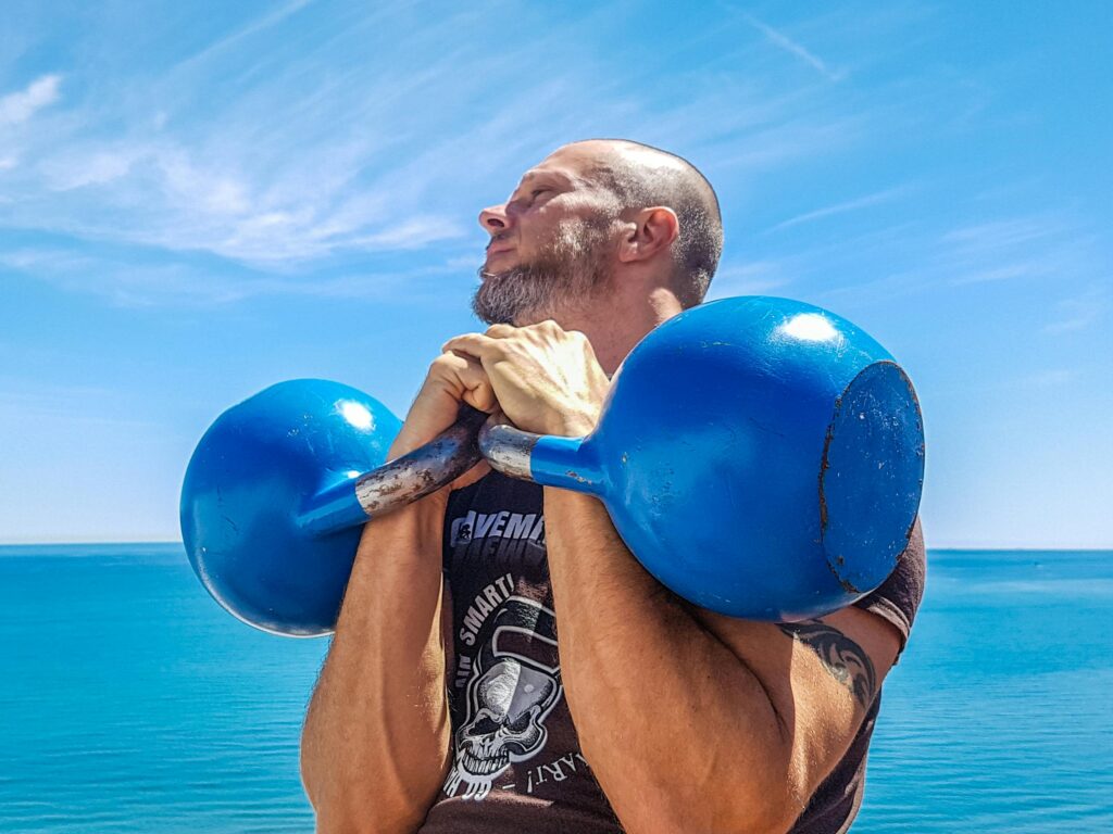 Athletic man lifting blue kettlebells by the ocean on a sunny day, showcasing strength and fitness.