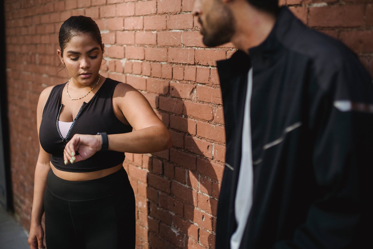 Two adults in sportswear, with one checking a smartwatch against a brick wall.