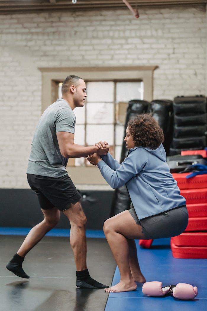 A personal trainer helps a client up in a fitness studio, surrounded by workout equipment.